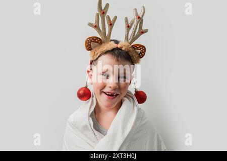 Un jeune garçon joyeux portant un bandeau de renne et orné de boules de Noël rouges sourit brillamment sur un fond Uni, incarnant la fête Banque D'Images