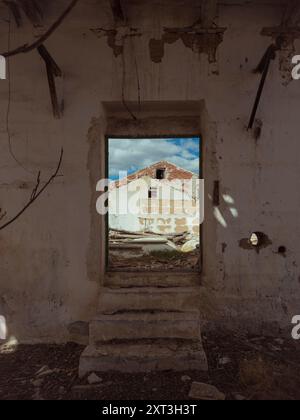 Cette image envoûtante capture une vue à travers une porte en décomposition, mettant en scène l'intérieur en ruine d'un bâtiment abandonné avec un ciel clair visible. Banque D'Images
