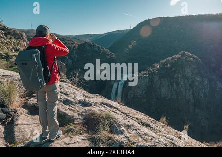 Une femme randonneuse se tient sur une corniche rocheuse, observant la majestueuse cascade Pozo de los Humos à Salamanque, enveloppée par un paysage accidenté Banque D'Images