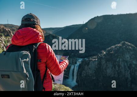 Une femme randonneuse dans une veste rouge vif se dresse à un point de vue, regardant la magnifique cascade Pozo de los Humos à Salamanque, profitant de la sérénité an Banque D'Images