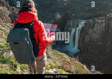 Une randonneuse en veste rouge observe la majestueuse cascade Pozo de los Humos à Salamanque, en Espagne, avec une carte à la main alors qu'elle se tient au milieu de gre luxuriant Banque D'Images