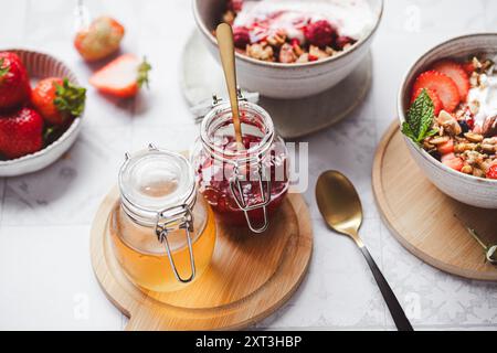 Petit déjeuner d'été sain avec bols de yaourt garni de granola, fraises fraîches et un brin de menthe. A proximité, bocaux de miel et Banque D'Images