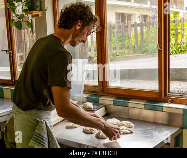 Un homme pétrissant la pâte à pizza et la pesant sur la balance sur une table en acier inoxydable, brillamment éclairé par la lumière naturelle d'une fenêtre donnant sur un gar luxuriant Banque D'Images