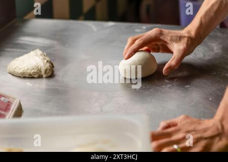 Une image détaillée capturant les mains d'une personne alors qu'elle pétrit habilement la pâte à pizza sur une surface métallique de cuisine, mettant en évidence le processus de fabrication de pizza Banque D'Images