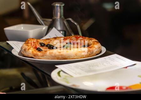 Pizza délicieusement cuite garnie d'olives noires et d'huile de basilic fraîche, servie sur une assiette en céramique accompagnée de condiments, créant un culinaire tentant Banque D'Images