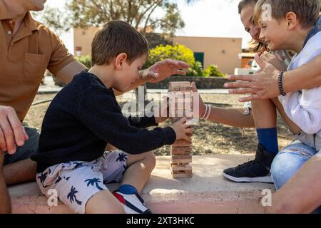 Cette image captivante capture une famille jouant à un jeu de jenga en bois dans le parc pendant l'automne. Une mère et un père, ainsi que leurs deux fils, le sont Banque D'Images