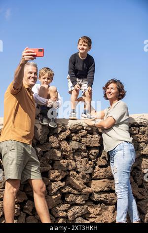Famille de cinq personnes, dont un père, une mère et trois fils, profite d'une journée ensoleillée en plein air alors qu'ils posent pour un selfie au sommet d'un mur de pierre rustique avec père, Banque D'Images