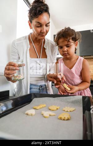 Une mère et sa jeune fille se livrent à la cuisson de biscuits, la mère saupoudrant des garnitures d'un pot pendant que l'enfant décore un cookie sur un four s. Banque D'Images