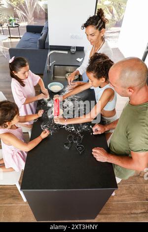 Une famille diversifiée, impliquant deux adultes et trois enfants, cuit avec enthousiasme des biscuits à la maison ils utilisent un rouleau à pâtisserie et emporte-pièces, sur Banque D'Images