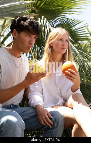 Un jeune couple se détend à l'ombre des palmiers, partageant un moment d'intimité et de rafraîchissement avec des fruits tropicaux leur tenue d'été décontractée com Banque D'Images