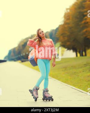 Jeune femme blonde élégante sur patins à roulettes avec sac à dos posant dans le parc de la ville Banque D'Images