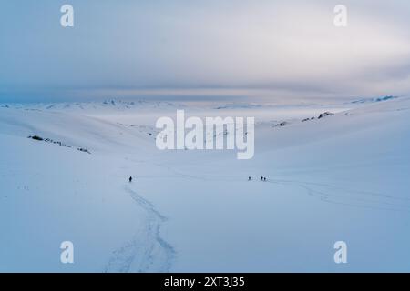 Des personnages méconnaissables traversent un immense paysage enneigé sous un vaste ciel au Kirghizistan, leurs chemins marqués par des empreintes de pas dans la neige immaculée. Banque D'Images