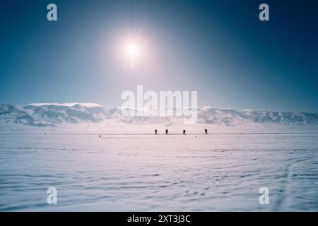 Des personnages méconnaissables voyagent à travers un vaste paysage enneigé sous un soleil éclatant au Kirghizistan, avec de majestueuses montagnes enneigées au loin. Banque D'Images