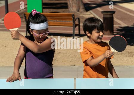 Deux enfants multiethniques pris dans un jeu de tennis de table ludique devant le garçon dans une chemise orange sourit pendant qu'il joue, tandis que la fille porte un violet Banque D'Images