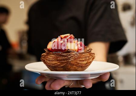 Serveur méconnaissable servant une délicieuse pâtisserie garnie de fruits frais, présentée sur une assiette blanche au fond flou. Banque D'Images