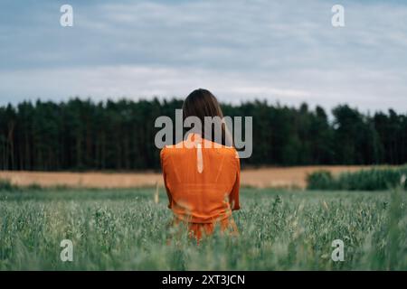 Vue arrière d'une femme brune méconnaissable dans une robe orange regarde loin vers une forêt lointaine, capturée de derrière au milieu d'un champ de hautes herbes. Banque D'Images