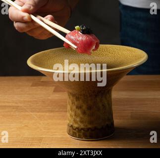 Un chef saisit un morceau de thon nigiri garni de caviar à l'aide de baguettes, planant au-dessus d'un bol en céramique texturé Banque D'Images