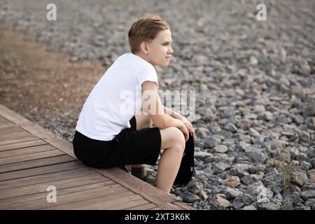 Adolescent assis seul sur un chemin en bois, regardant loin réfléchi, portant un t-shirt blanc et un short noir, au milieu d'un fond de gravier. Banque D'Images