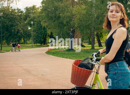 Jeune femme avec vélo dans l'allée. Belle femme en vêtements décontractés debout avec vélo avec panier. Femme active dans un parc public. Banque D'Images