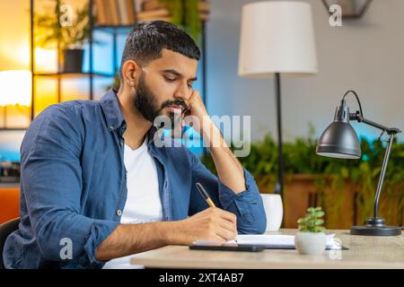 Homme barbu indien freelance travaillant avec des documents du bureau à domicile, analysant des informations, lisant des documents d'affaires préparent le rapport financier. Jeune homme hispanique comptable analysant des documents graphiques. Banque D'Images