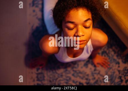 Un Africain attrayant pratique le yoga du soir. Une charmante fille noire bouclée dans un costume de fitness étire son dos sur le sol dans le salon. Haut vi Banque D'Images