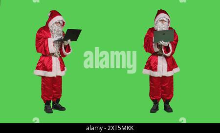 Santa claus cosplay fonctionne avec un ordinateur portable sur fond de corps plein écran vert, en utilisant le navigateur en ligne en studio. Père noël recherche le site Internet alors qu'il est habillé avec costume et chapeau. Banque D'Images