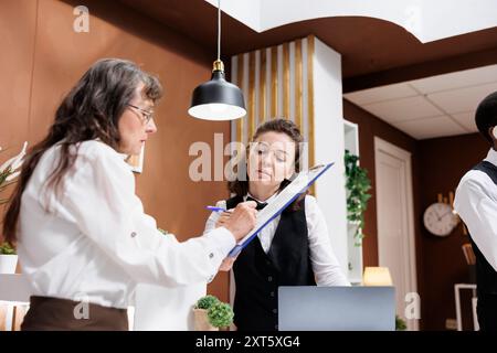 Une femme âgée retraitée arrive à la réception de l'hôtel et remplit le formulaire de réservation, souriant et parlant avec la réceptionniste. Touriste âgée signant les documents d'enregistrement à la réception. Banque D'Images