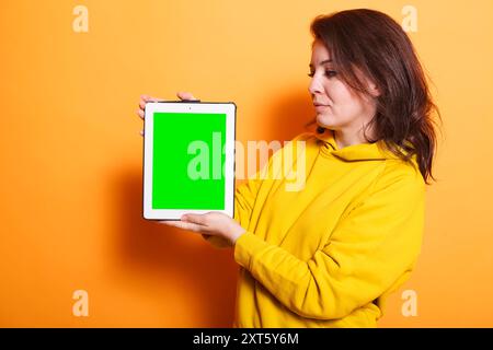Femme caucasienne saisissant verticalement la tablette avec l'écran vert vers la caméra. Portrait d'une personne féminine moderne debout et tenant un appareil numérique avec un modèle de maquette chromakey vierge. Banque D'Images