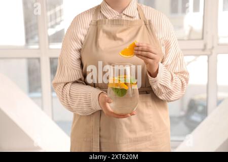 Jeune femme tenant une cruche d'eau infusée avec de l'orange et de la menthe près de la fenêtre à la maison Banque D'Images