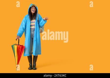 Heureuse jeune femme afro-américaine en imperméable avec parapluie sur fond jaune Banque D'Images