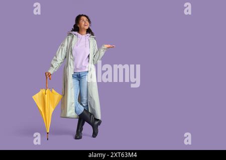 Heureuse jeune femme afro-américaine en imperméable avec parapluie sur fond lilas Banque D'Images