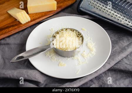 Fromage parmesan finement râpé dans une tasse à mesurer en métal : fromage parmesan reggiano fraîchement râpé avec un coin de fromage et une râpe en arrière-plan Banque D'Images