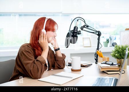 Une belle et positive podcaster féminine asiatique avec les cheveux bouclés rouges, portant des écouteurs, enregistre son podcast dans le studio, parlant dans le micro Banque D'Images