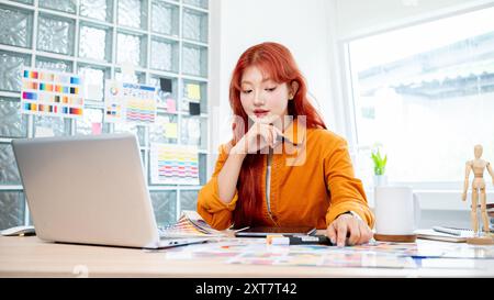Une graphiste féminine asiatique belle et créative avec les cheveux bouclés rouges travaille dans son studio de création, se concentrant sur la sélection des couleurs d'un ensemble Banque D'Images