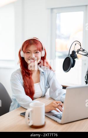 Une charmante femme asiatique podcaster ou créatrice de contenu aux cheveux roux porte des écouteurs, utilise son ordinateur et sourit à la caméra en étant assise Banque D'Images