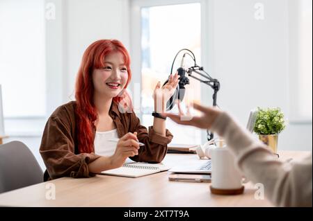 Une podcastrice asiatique confiante et amicale ou une créatrice de contenu aux cheveux bouclés rouges interviewe une invitée dans son studio, profitant de la conversation. Banque D'Images