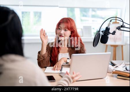 Une podcasteuse asiatique confiante et attrayante aux cheveux bouclés rouges interviewe une invitée, enregistrant son nouvel épisode de podcast en studio. Banque D'Images