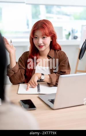 Une podcasteuse asiatique confiante et attrayante aux cheveux bouclés rouges interviewe une invitée, enregistrant son nouvel épisode de podcast en studio. Banque D'Images