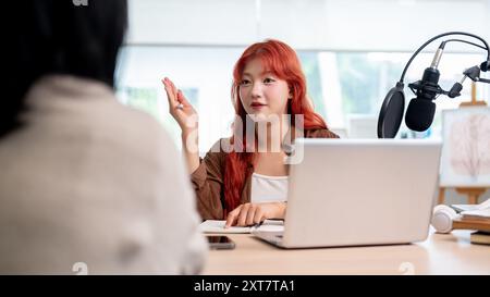 Une podcasteuse asiatique confiante et attrayante aux cheveux bouclés rouges interviewe une invitée, enregistrant son nouvel épisode de podcast en studio. Banque D'Images