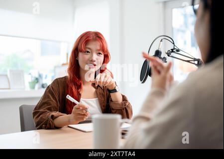 Une podcasteuse asiatique confiante et attrayante aux cheveux bouclés rouges interviewe une invitée, enregistrant son nouvel épisode de podcast en studio. Banque D'Images