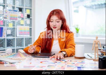 Une graphiste féminine asiatique belle et créative avec les cheveux bouclés rouges travaille à son bureau dans le studio, sélectionnant des couleurs à partir d'un ensemble de couleurs Banque D'Images