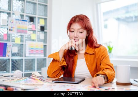 Une graphiste féminine asiatique belle et créative avec les cheveux bouclés rouges travaille à son bureau dans le studio, sélectionnant des couleurs à partir d'un ensemble de couleurs Banque D'Images