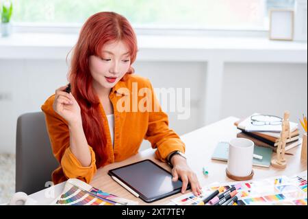 Une graphiste asiatique créative et concentrée avec les cheveux roux travaille à son bureau dans le studio, conçoit un nouveau projet et sélectionne des couleurs Banque D'Images
