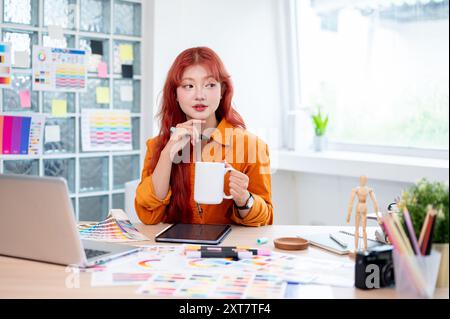 Une graphiste asiatique créative et attrayante avec des cheveux bouclés rouges tient une tasse à café, regardant loin réfléchi, reposant son menton sur son h. Banque D'Images