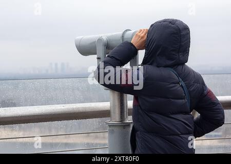 Un touriste regarde à travers un télescope un panorama de la ville par temps pluvieux Banque D'Images