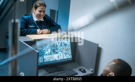 Afro-américain travailleur de la sécurité masculin assis sur le lieu de travail avec un moniteur d'ordinateur montrant des caméras de surveillance vidéo, utilise un téléphone portable. Garde féminine parle à un collègue, donne les clés. Système de vidéosurveillance. Banque D'Images