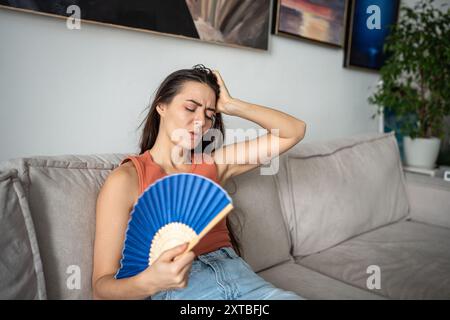 Femme surchauffée soufflant avec ventilateur de papier, assise sur le canapé à la maison, essayant de refroidir à la maison Banque D'Images