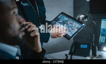Une employée montre des images de vidéosurveillance à un agent de sécurité afro-américain à l'aide d'une tablette. Divers opérateurs de sécurité contrôlent les caméras de surveillance sur les écrans d'ordinateur dans le centre de surveillance. Banque D'Images