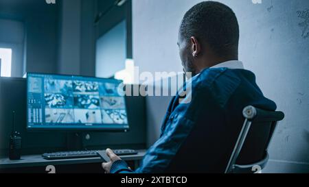 L'agent de sécurité masculin afro-américain utilise un téléphone portable assis sur le lieu de travail dans le bureau. Moniteur d'ordinateur avec affichage en temps réel des caméras de surveillance. Système de vidéosurveillance et sécurité sociale. Banque D'Images