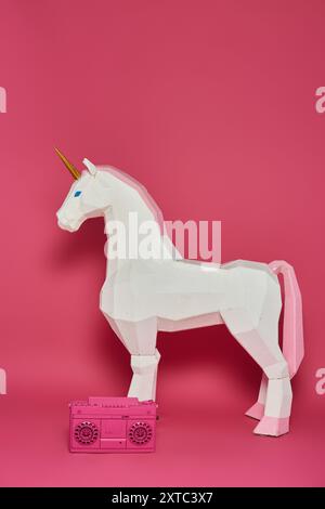 Une licorne en origami blanc se tient devant un fond rose avec une boombox rose à ses pieds. Banque D'Images